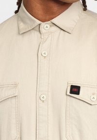 Beige Hemd mit Knopfleiste, spitzen Kragen, zwei Brusttaschen und einem kleinen roten Emblem auf der linken Tasche. Weicher Stoff.