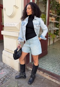 Ljusblå denimjacka, svart polotröja, ljusa shorts, svarta knähöga stövlar med spännen, håller en svart vävd handväska.