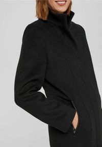 Manteau en mélange de laine noir avec col haut, coupe slim et poches latérales zippées. Présente une texture lisse et un design minimaliste sans motifs.