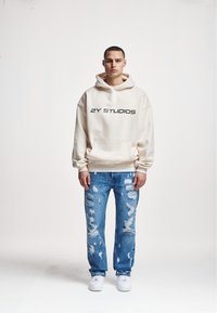 Beige hoodie with front pocket and black "2Y STUDIOS" text, paired with light blue distressed jeans and white sneakers. Simple, casual design.
