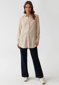 Hellbeige Hemd mit langen Ärmeln und Knöpfen, verziert mit hellblauen Blumenmustern, kombiniert mit dunkelblauen weit geschnittenen Hosen und Turnschuhen.