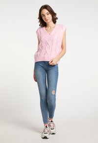 Roze gebreide mouwloze trui met een V-hals, met een gestructureerd kabelontwerp, gecombineerd met blauwe skinny jeans en meerkleurige sneakers.