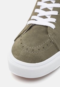 Olivengrøn ruskinds sneakers med hvide snørebånd, perforeret tåområde og en tekstureret hvid gummisål. Nærbillede af skoens front.