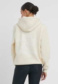 Krämfärgad hoodie med en stor vit grafisk text som säger "DET SOM GÅR RUNT KOMMER TILLBAKA", med en kenguruficka och huva.