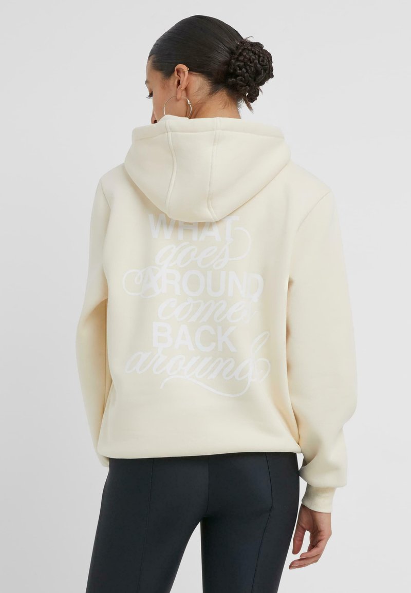 Krämfärgad hoodie med en stor vit grafisk text som säger "DET SOM GÅR RUNT KOMMER TILLBAKA", med en kenguruficka och huva.