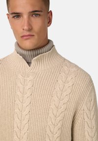 Beige stickad tröja med hög ribbad krage. Har ett kabelmönster framtill och en strukturerad design genom hela tröjan.