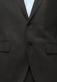 Σκούρο γκρι σακάκι κοστουμιού με δύο μαύρα κουμπιά και τσέπες με καπάκι πάνω από ένα ανοιχτό μπλε πουκάμισο.