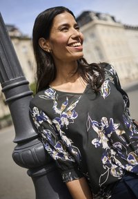 Blusa nera con stampa floreale nei toni del blu, bianco e oro. Presenta un collo a V e maniche arricciate. Texture liscia su uno sfondo di pietra.
