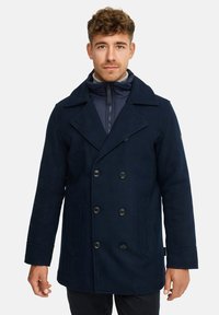 Marineblå uldblandingspeacoat med dobbeltbrystet front, store kraver og to sidelommer. Har sorte knapper og et lynlåslukket indvendigt lag.