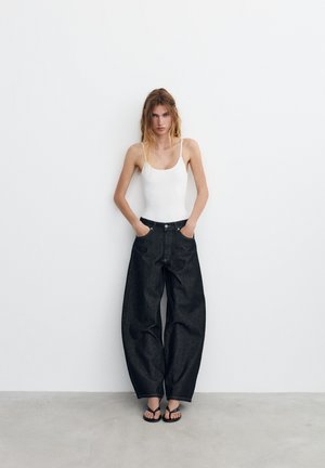 Junge Frau mit langen Haaren steht vor einer weißen Wand, trägt ein weißes Tanktop, schwarze weite Jeans und schwarze Sandalen, die Hände in den Taschen.