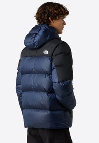 Puffjacka i blått och svart, med huva, quiltat mönster, dragkedjefickor och logotyp-detalj på övre ryggen. Slät textur.