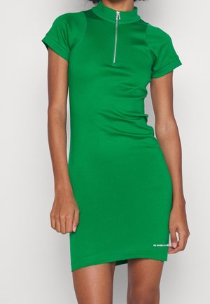 Femme portant une robe verte ajustée à manches courtes avec une petite fermeture éclair frontale et un logo discret près de l'ourlet.