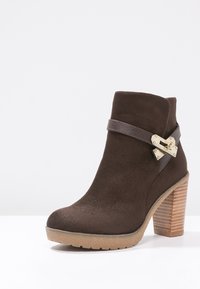 Brun mocka ankelboot med rund tå, ett läderrem och guldhardwaredetaljer, samt en texturerad träklack.
