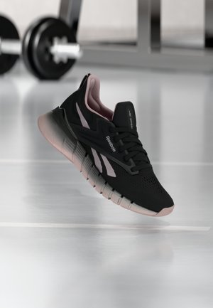 Black and pink Reebok running shoe with cushioned sole floating above a gym floor with a blurred dumbbell in the background.