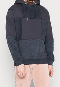 Felpa blu navy con pannelli di varia texture; presenta un cappuccio con cordoncino e polsini a costine, progettata con dettagli di cucitura in evidenza.