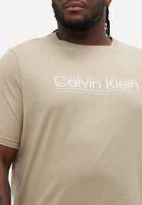 Bež majica s kružnim ovratnikom napravljena od mekog materijala, s bijelim natpisom "Calvin Klein" i "OSNOVANO TISUĆU DEVETSTO ŠEZDESET I OK" ispod njega.