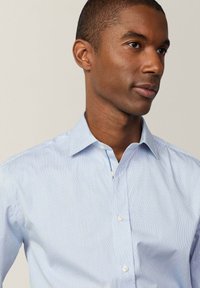 Camisa de vestir a rayas de color azul claro, con cuello puntiagudo, cierre frontal con botones y una textura de tela suave. Presenta detalles sutiles en contraste.