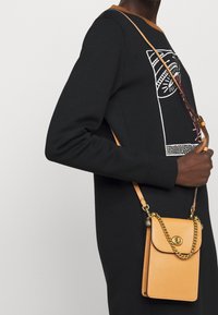 Läderväska i tanfärg med texturerad yta, guld kedjesnor och magnetisk stängning, bärs med en svart sweatshirt med grafisk design.