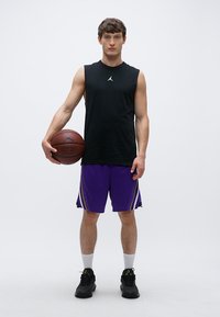 Tricou negru fără mâneci, pantaloni scurți mov cu dungi galbene, ținând un coș de baschet, pantofi negri, stând pe un fundal neutru.