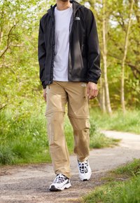 Veste légère noire portée sur une chemise blanche, associée à un pantalon cargo beige. Chaussures de sport noires et blanches. Environnement naturel vert.