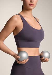 Ljubičasti sportski grudnjak s širokim naramenicama i glatkom teksturom. Model drži dvije metalik srebrne vježbačke lopte. Neutralna pozadina.