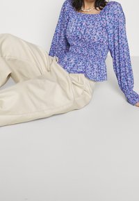 Langärmlige Peplum-Bluse mit floralen Mustern in Blau-, Rosa- und lila Farbtönen; elastischer Taillen- und Halsausschnitt; kombiniert mit hellbeigen Hosen.