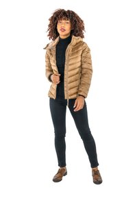 Femme aux cheveux bouclés portant une veste matelassée beige par-dessus un col roulé noir, un pantalon noir et des chaussures à lacets marron, se tenant devant un fond blanc.