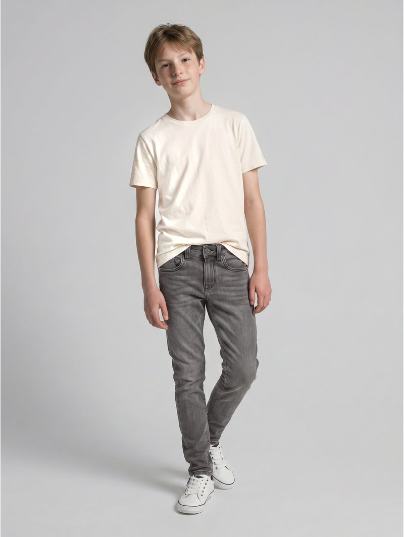 Jongen staat met een effen lichtbeige T-shirt, grijze spijkerbroek en witte sneakers tegen een effen lichtgrijze achtergrond.