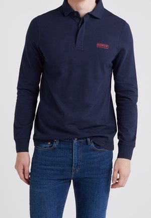 Man wearing a navy long-sleeve polo shirt with a small red Barbour International logo on the chest and blue jeans.
