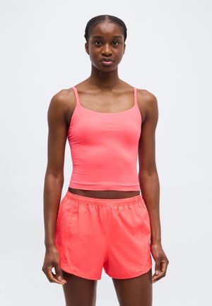 Žena v jasne ružovom priliehavom tielku a zodpovedajúcich elastických šortkách s vysokým pásom, otočená čelom k svetlému jednoliatemu pozadiu.