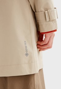 Beżowa kurtka Gore-Tex o gładkiej teksturze. Posiada mankiety u rękawów, widoczne przeszycia oraz logo Gore-Tex z lewej strony.