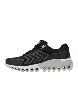 Chaussure de sport noire avec motif chevron gris, semelle blanche avec découpes circulaires et panneau de maintien de talon gris.