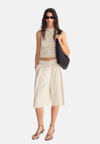 Top corto sin mangas de color blanco con un patrón texturizado; culottes beige con plisados; tacones de tiras; bolso de mano tejido en negro; gafas de sol.