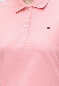 Polo rose à col avec cinq boutons et un petit logo brodé rouge, blanc et bleu sur le côté gauche de la poitrine.