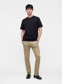 Junger Mann steht mit den Händen in den Taschen, trägt ein schwarzes T-Shirt, beige Hose und schwarze elegante Schuhe vor einem schlichten weißen Hintergrund.