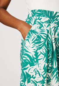 Spódnica w zielony wzór liści na białym tle, z bocznymi kieszeniami, usztywnionym pasem i lekko opadającym krojem. Gładka tekstura tkaniny.