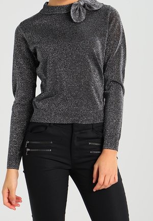 Femme portant un haut gris scintillant à manches longues avec un détail nœud sur le col et un pantalon noir avec poches zippées.