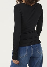 Woman wearing a fitted black long-sleeve top and blue denim jeans, shown from the back with one hand relaxed by her side.