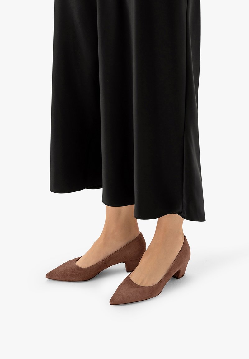 Jambes portant des chaussures marron à talons bas et bouts pointus, associées à un pantalon noir à jambes larges et courtes, sur un fond uni.