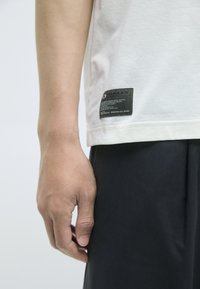 Hvit bomulls t-skjorte med korte ermer, med en mørk etikett ved kanten som viser merke og pleieinformasjon, parret med svarte shorts.