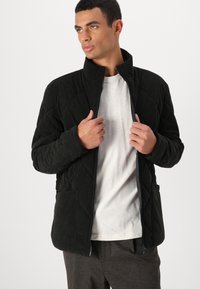 Μαύρο σακάκι καπλάνι με σχέδιο διαμαντιού, ψηλό γιακά, πλαϊνές τσέπες και μπροστινό φερμουάρ. Φοριέται πάνω από ένα ανοιχτό γκρι T-shirt.