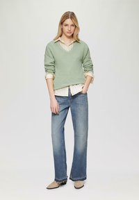 Svetlozeleni pulover z oblikovanim izrezom in teksturiranim materialom, nosen nad belim prugastim srajcem, v kombinaciji s svetlo modrimi širokimi jeans hlačami in bež čevlji s koničastim prstom.