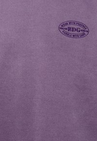 Lila bomull T-shirt med en texturerad yta som har en cirkulär lila logotyp med texten "BDG" och skötselråd.