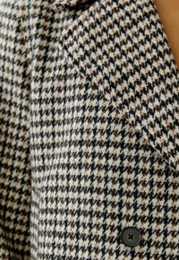 Nahaufnahme eines Blazers im schwarz-weißen und braunem Hahnentritt-Muster mit einem schwarzen Knopf und sichtbarem Revers.