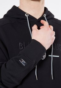 Haut à capuche noir avec une poche kangourou, présentant un texte brodé, une finition texturée et des cordons de serrage contrastants avec un motif en chevron.