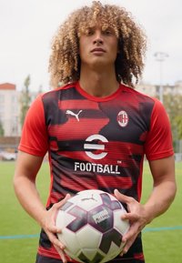 Camiseta de fútbol de manga corta en rojo y negro con un patrón, que presenta el logo de Puma y un emblema circular. El jugador sostiene un balón de fútbol en blanco y negro.