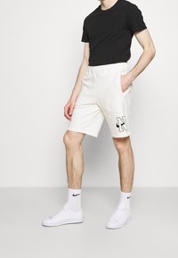 Vita shorts i bomull med elastisk midjeband, utrustade med sidofickor och en svart "N"-logotyp. Bärs med en svart t-shirt och vita sneakers.