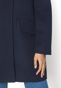 Mörkblå ullkappa med ärm och ficka, där en persons hand vilar bredvid, som bär blå denimjeans.