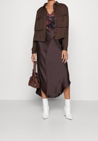 Chemise en denim marron à manches courtes sur un haut violet à motifs, associée à une jupe portefeuille en satin marron et des bottines blanches à talons aiguilles. Sac marron.