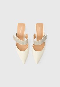 Λευκές γόβες με μυτερή μύτη και λουράκι πίσω από το τακούνι, διακοσμημένο με ασημένιο λουρί με στρας. Λείο δερμάτινο εξωτερικό και μπεζ πάτος.